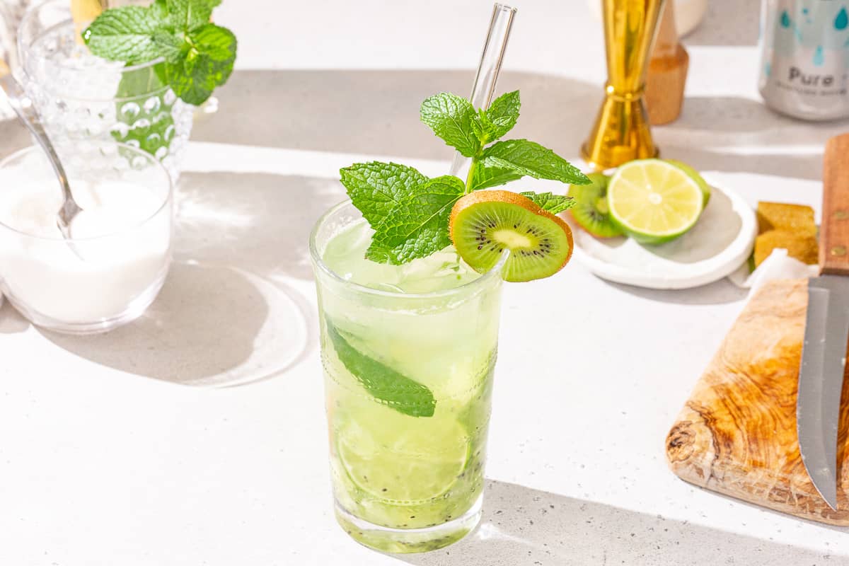 Overhead view of Kiwi Mojito in a glass. The drink is garnished with a kiwi slice, mint sprig and lime slice, and has a straw. In the background a cut kiwi and lime are visible along with a gold jigger. On the left is an extra mint sprig.