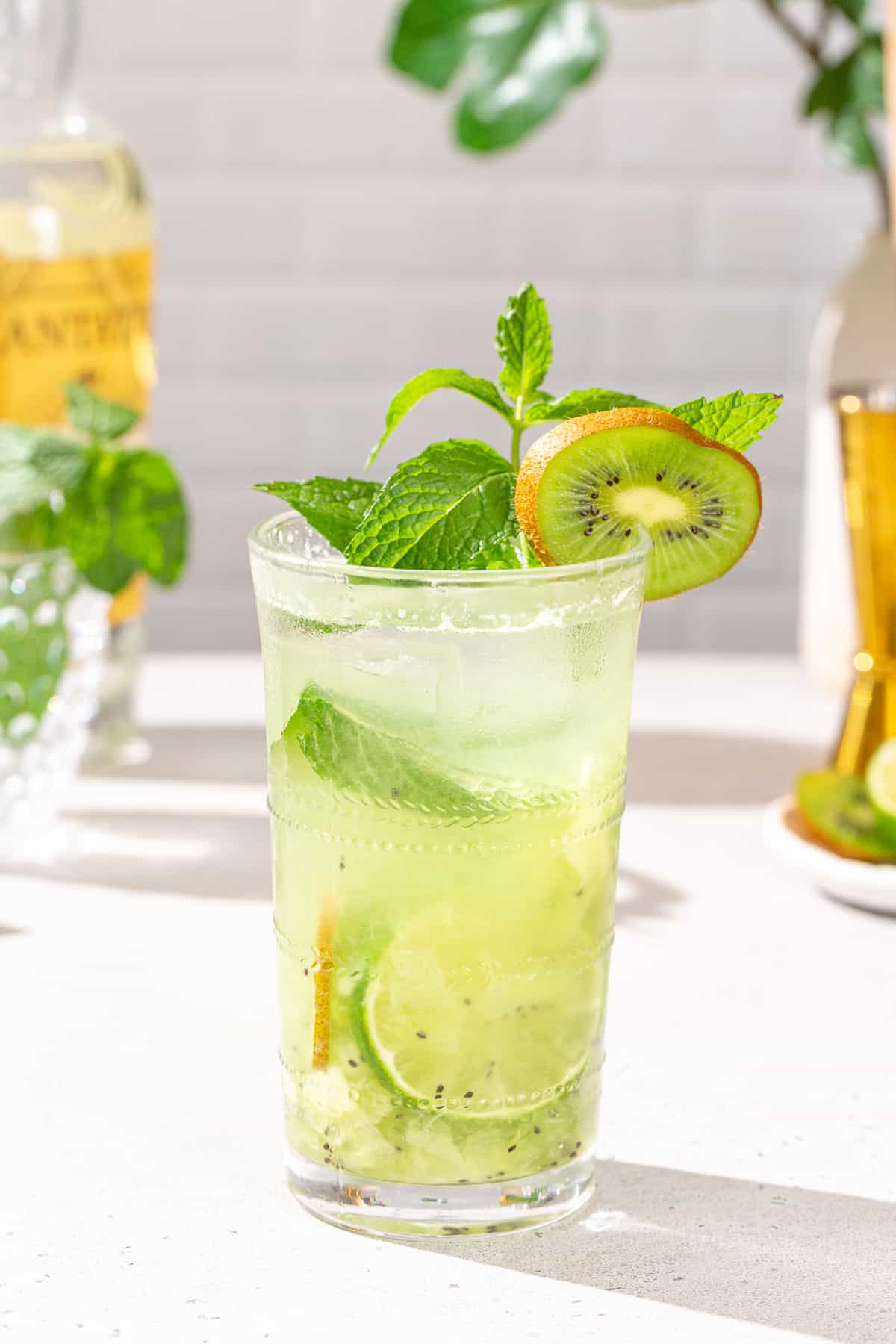 Side view of Kiwi Mojito cocktail in a tall glass. The drink is garnished with a kiwi slice, mint sprig and lime slice. In the background are ingredients used in the drink, including rum, mint and lime, as well as a jigger.