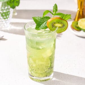 Overhead view of Kiwi Mojito in a glass. The drink is garnished with a kiwi slice, mint sprig and lime slice, and has a straw. A cut kiwi and lime are in the background.
