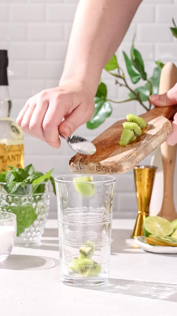 Hand using a spoon to add fresh kiwi pieces to a large cocktail glass.