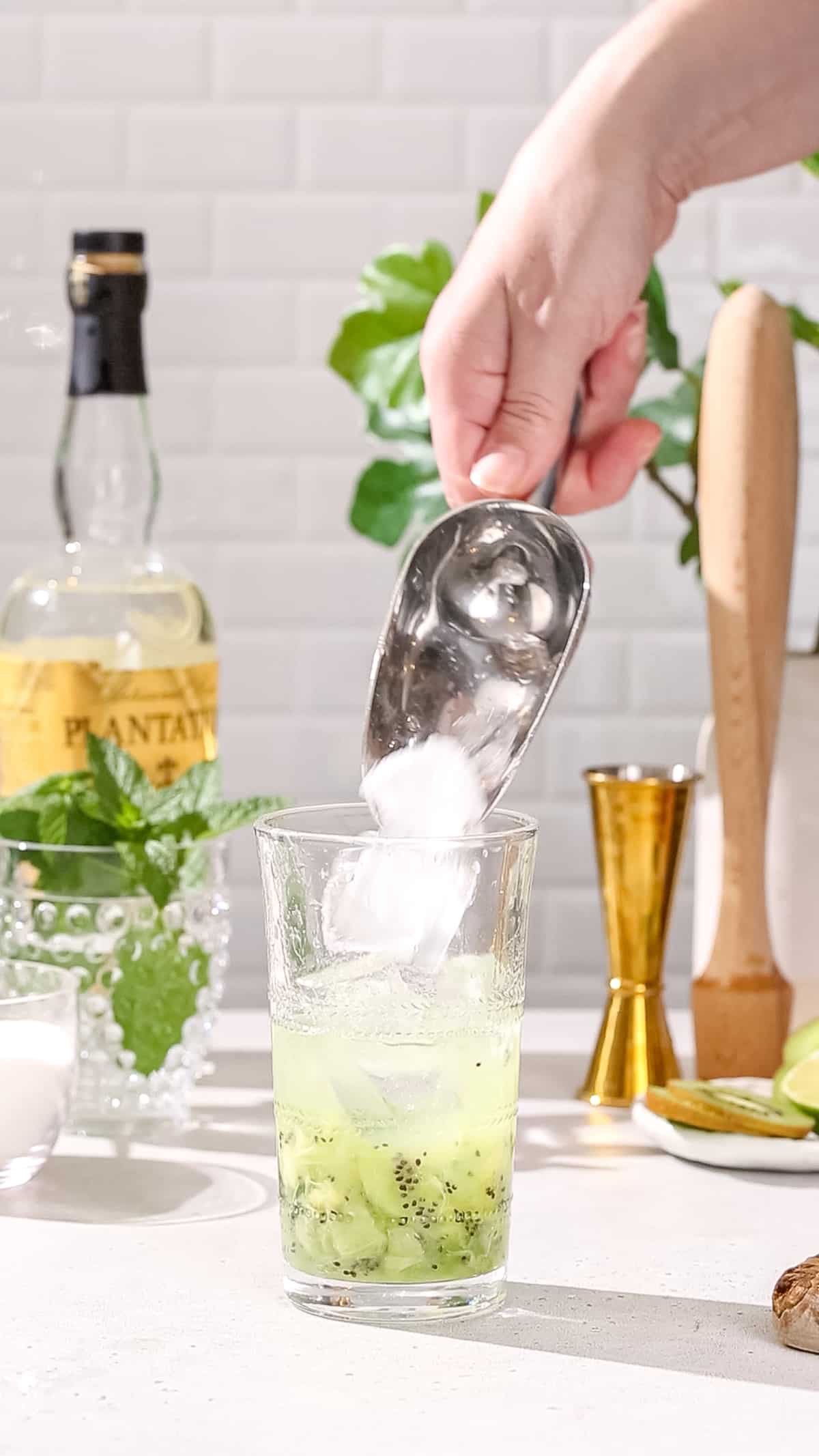 Hand using an ice scoop to add ice to a cocktail glass with kiwi and lime juice and rum in it.
