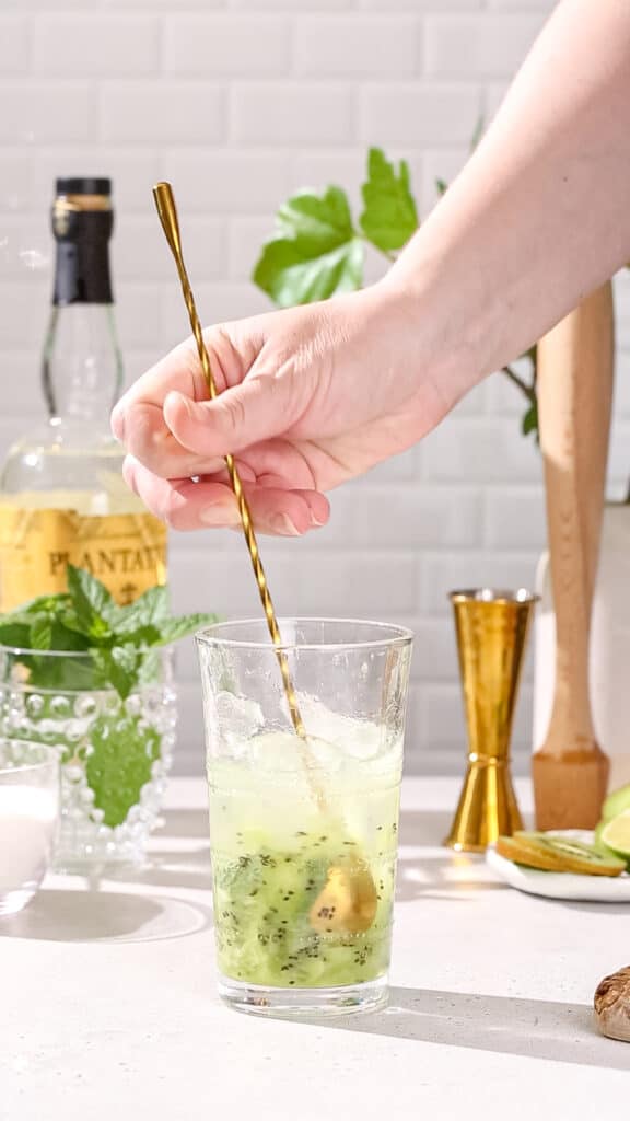 Hand using a long bar spoon to stir a mojito cocktail.