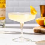 Side view of an Orange Daiquiri cocktail in a coupe glass with an orange peel garnish. In the background are a cut orange and lime on a cutting board, a gold jigger, and a bottle of white rum.