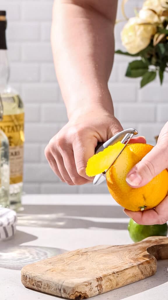 Hands using a vegetable peeler to peel an orange.