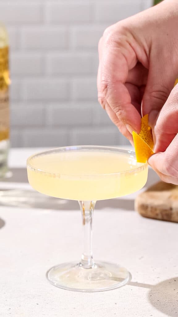 Hands adding an orange peel to the side of a coupe glass for a garnish.
