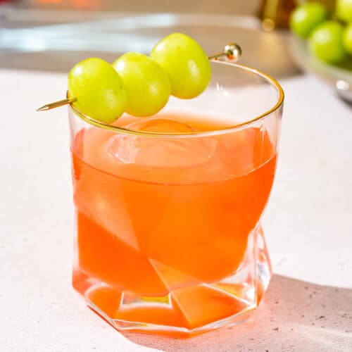 Close up detailed view of an Enzoni cocktail in a gold-rimmed old fashioned cocktail glass. The reddish-orange colored drink is garnished with three green grapes on a cocktail pick.