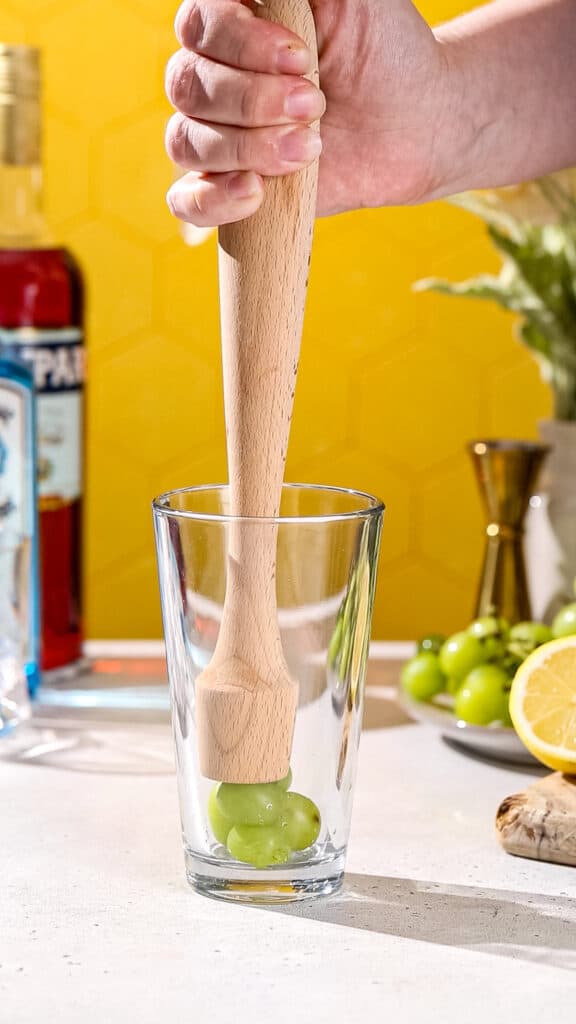 Hand using a wooden muddler to smash green grapes in a cocktail shaker.