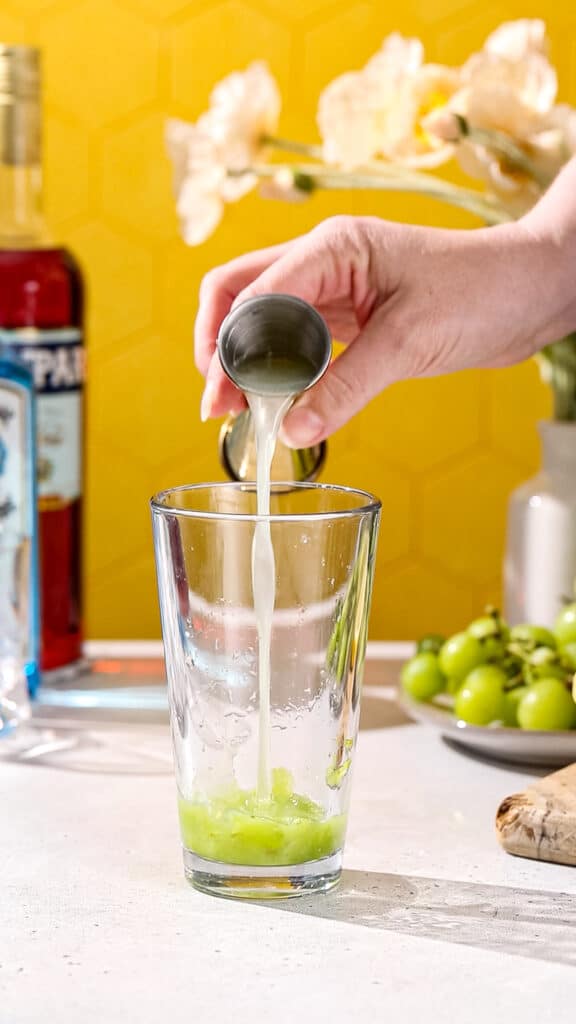 Hand pouring lemon juice into cocktail shaker that has smashed green grapes in it.