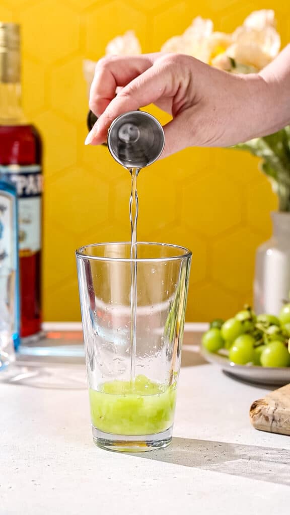 Hand pouring simple syrup into cocktail shaker using a gold jigger.