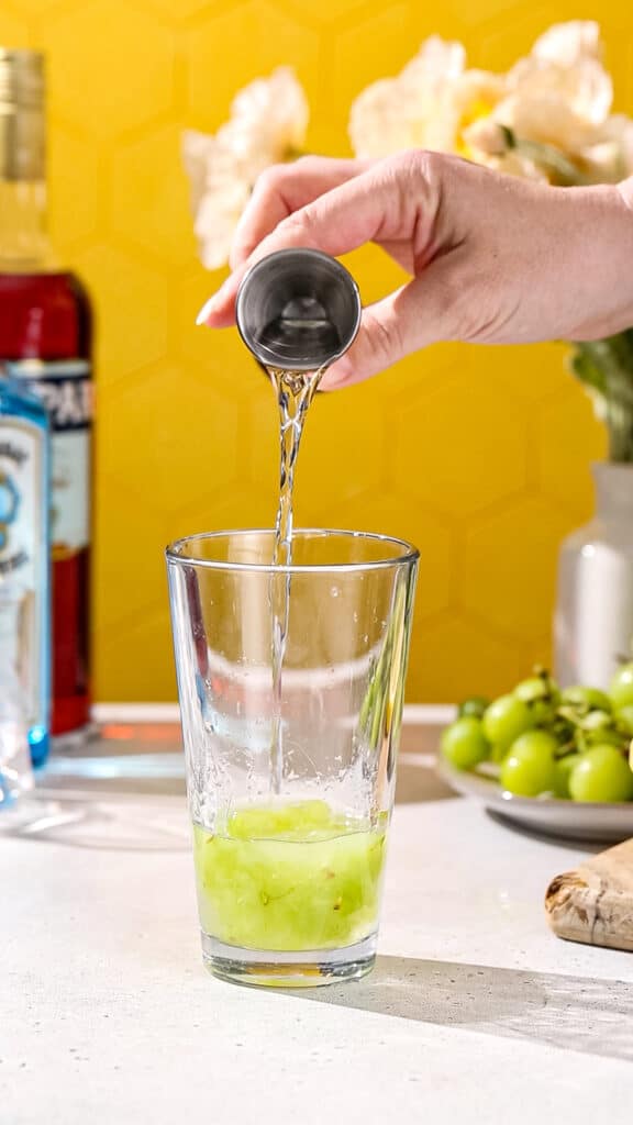 Hand pouring gin into a cocktail shaker filled with green liquid.