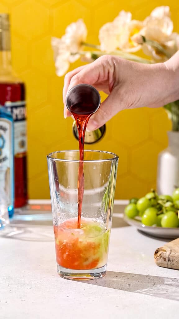 Hand pouring Campari liqueur into a cocktail shaker filled with green liquid.