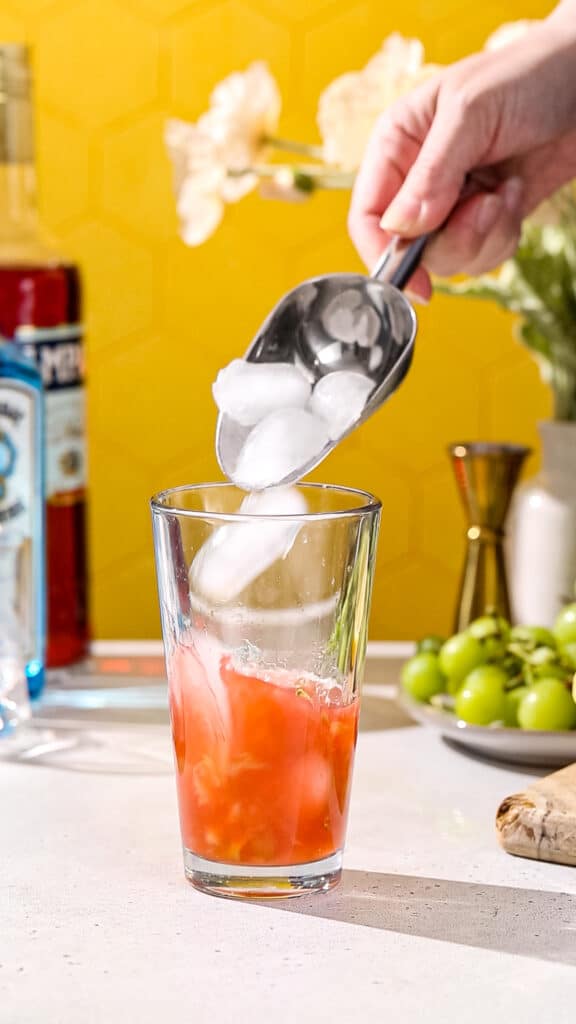 Hand using an ice scoop to add ice to a cocktail shaker filled with red liquid.