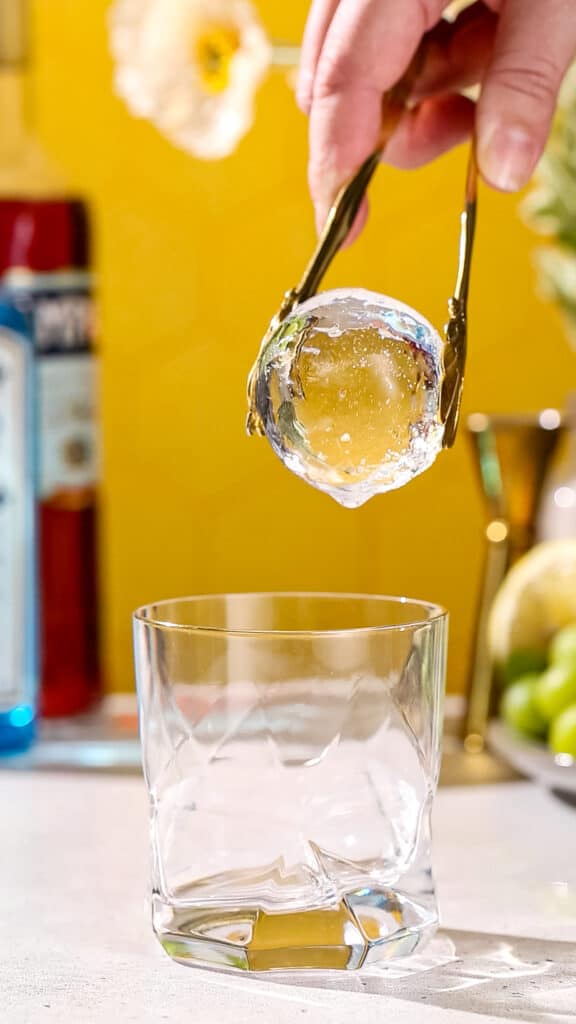Gold tongs adding a large ice sphere to an old fashioned cocktail glass.