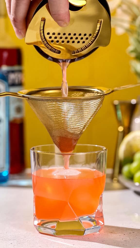 Red colored drink being double strained into a cocktail serving glass.