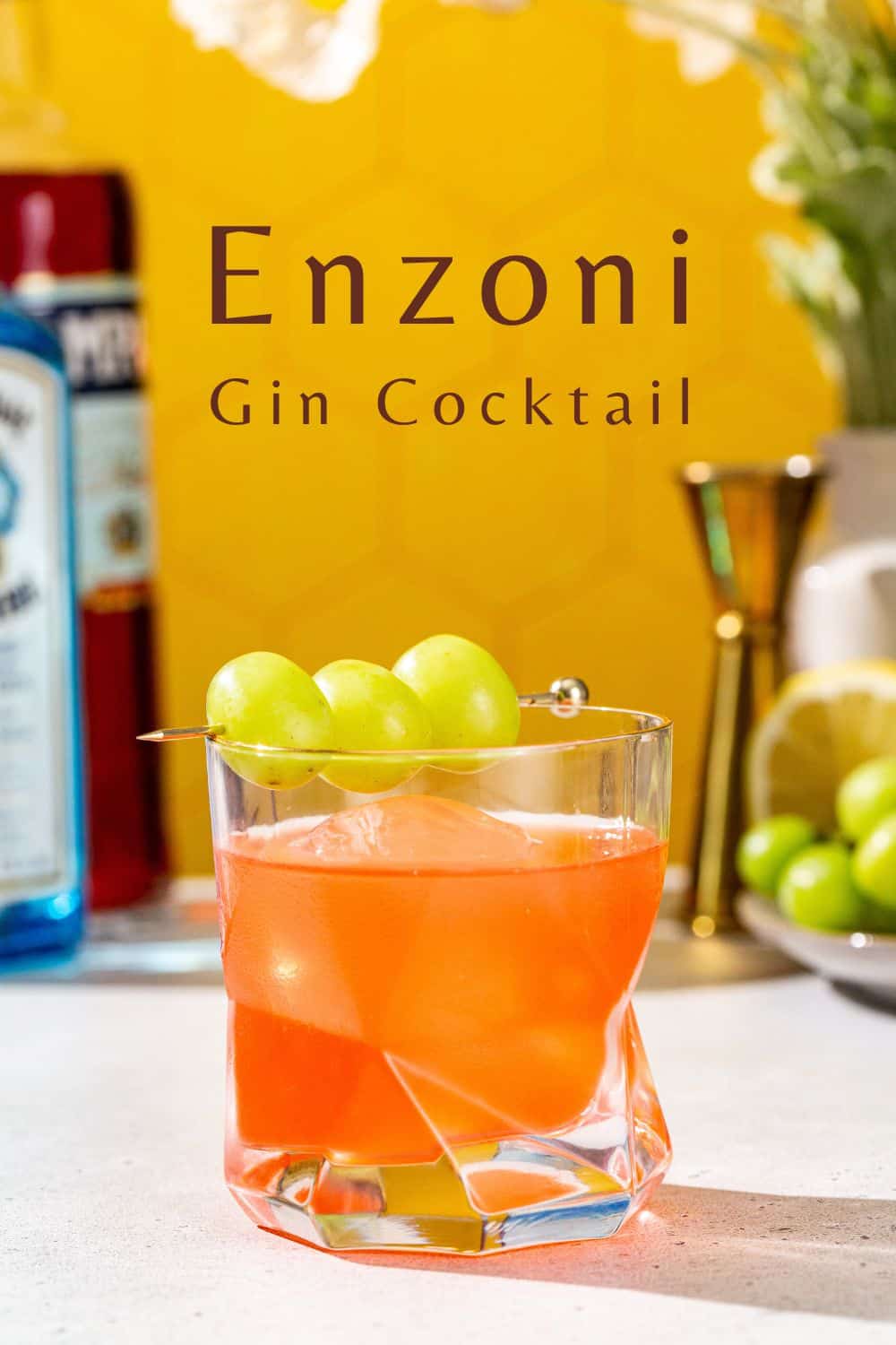 Side view of Enzoni cocktail in an old fashioned cocktail glass. In the background is a bottle of gin and Campari, along with a gold jigger, a cut lemon and green grapes. Text above the drink says “Enzoni gin cocktail”.