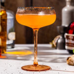 Side view of an orange colored Halloween Margarita in a coupe cocktail glass. The drink has a black salt rim and ingredients are visible in the background.