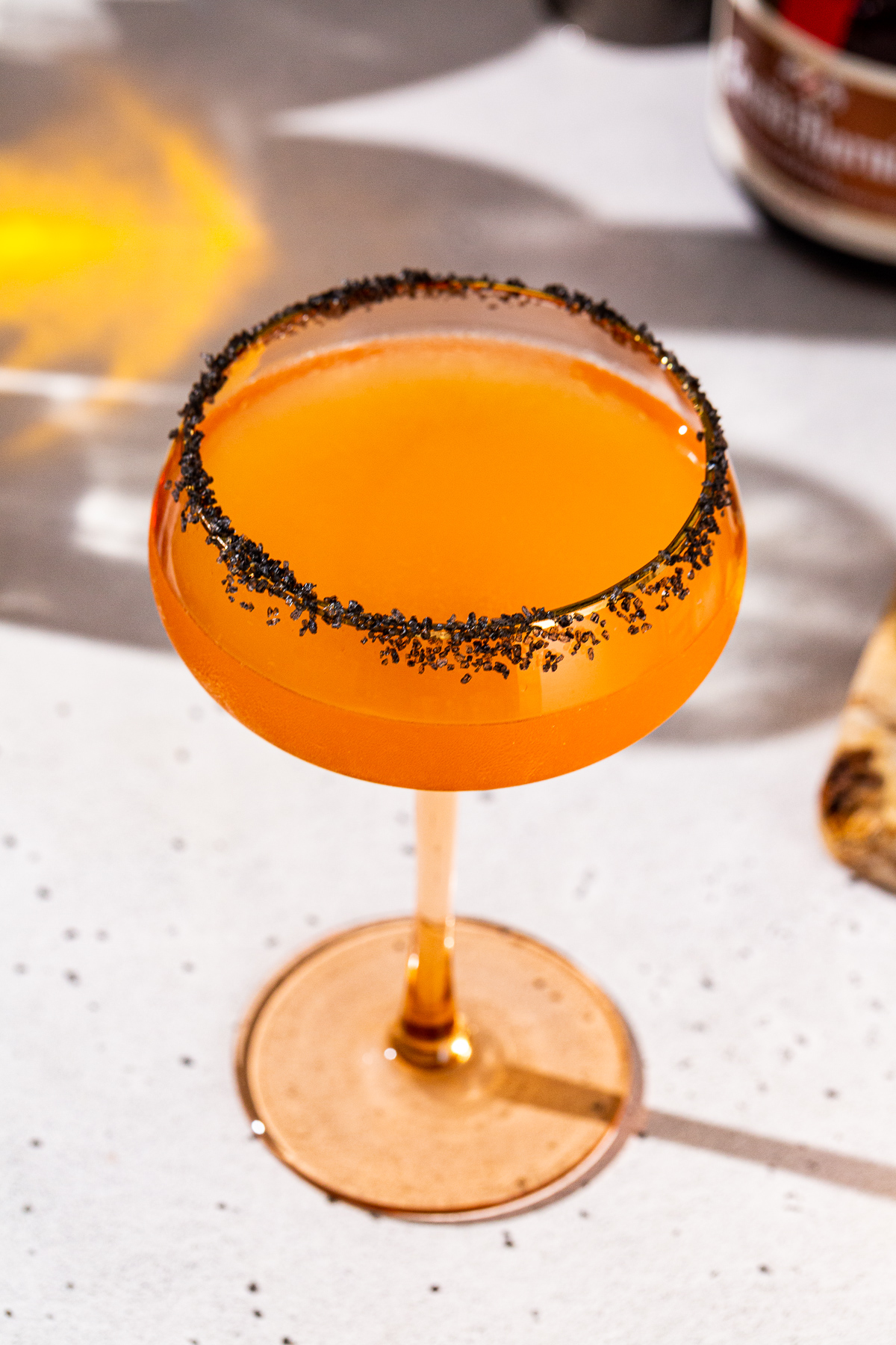 Overhead view of a Halloween Margarita. The drink is in an orange tinted coupe glass and has a black lava salt rim. Grand Marnier is seen in the background.