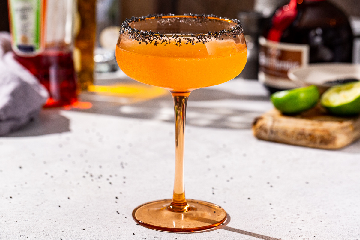 Side view of a Halloween Margarita with a black salt rim on a countertop. A Lime, a bottle of Grand Marnier, Licor 43 and tequila are all visible in the background.