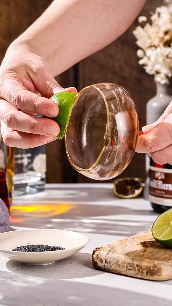 Hand using a cut lime to wet the rim of a coupe cocktail glass.