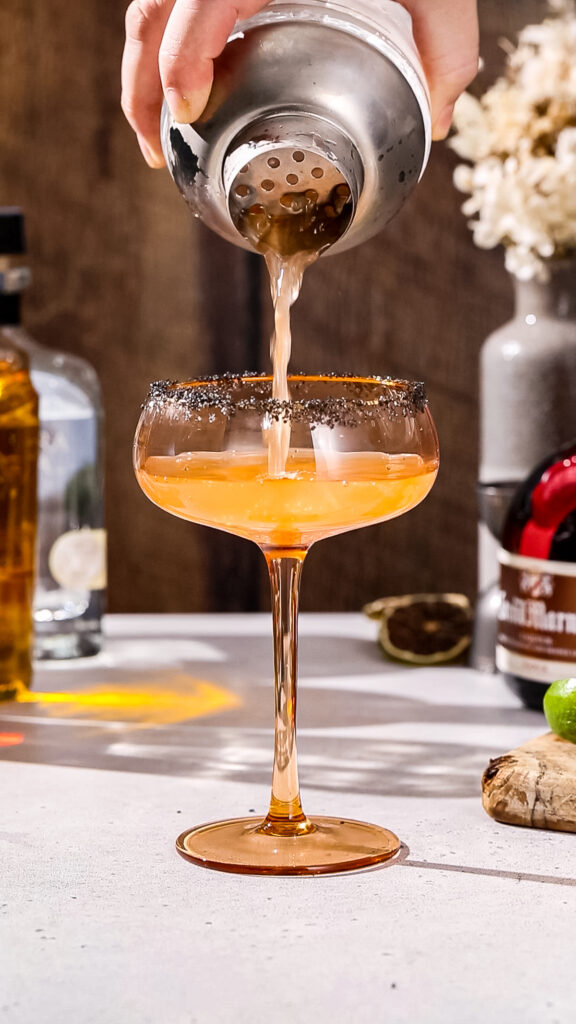 Hand pouring the cocktail into a coupe glass with a black salt rim.