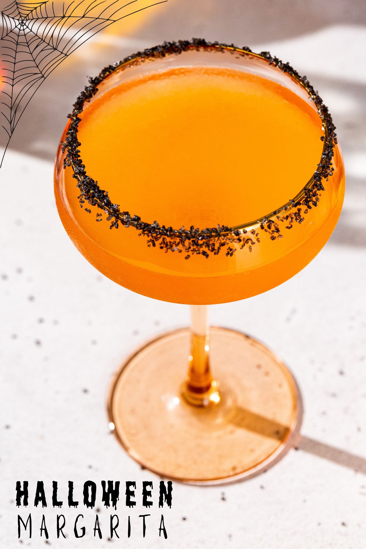 Overhead view of a Halloween Margarita. The drink is in an orange tinted coupe glass and has a black lava salt rim. Text at the bottom says Halloween Margarita.