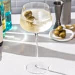 Close up of an Italicus Spritz cocktail in a large stemmed cocktail glass, with ingredients in the background. The drink has ice in it and looks refreshing. It’s bubbly and garnished with three green olives on a cocktail pick.
