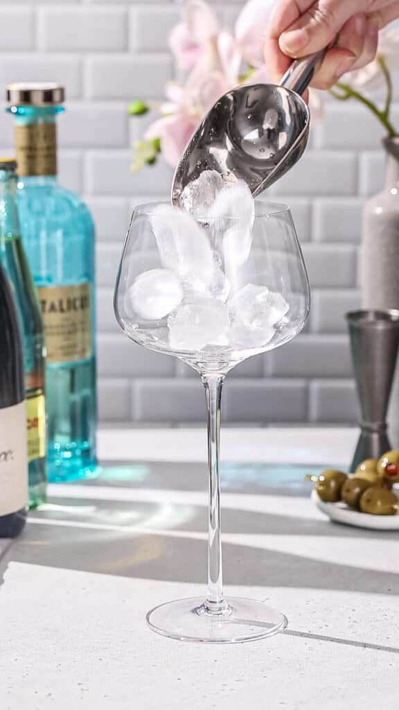 Hand using an ice scoop to add ice to a large stemmed cocktail glass.
