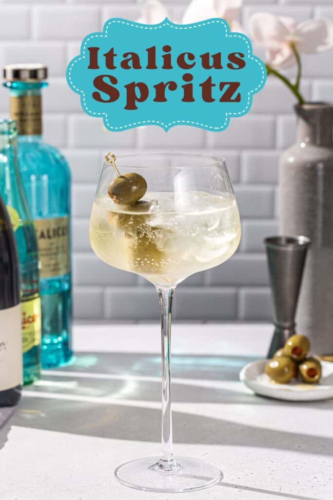 Italicus Spritz cocktail in a large stemmed cocktail glass, with ingredients in the background. The drink is bubbly and garnished with three green olives on a cocktail pick. Bold text above the drink says “Italicus Spritz”.
