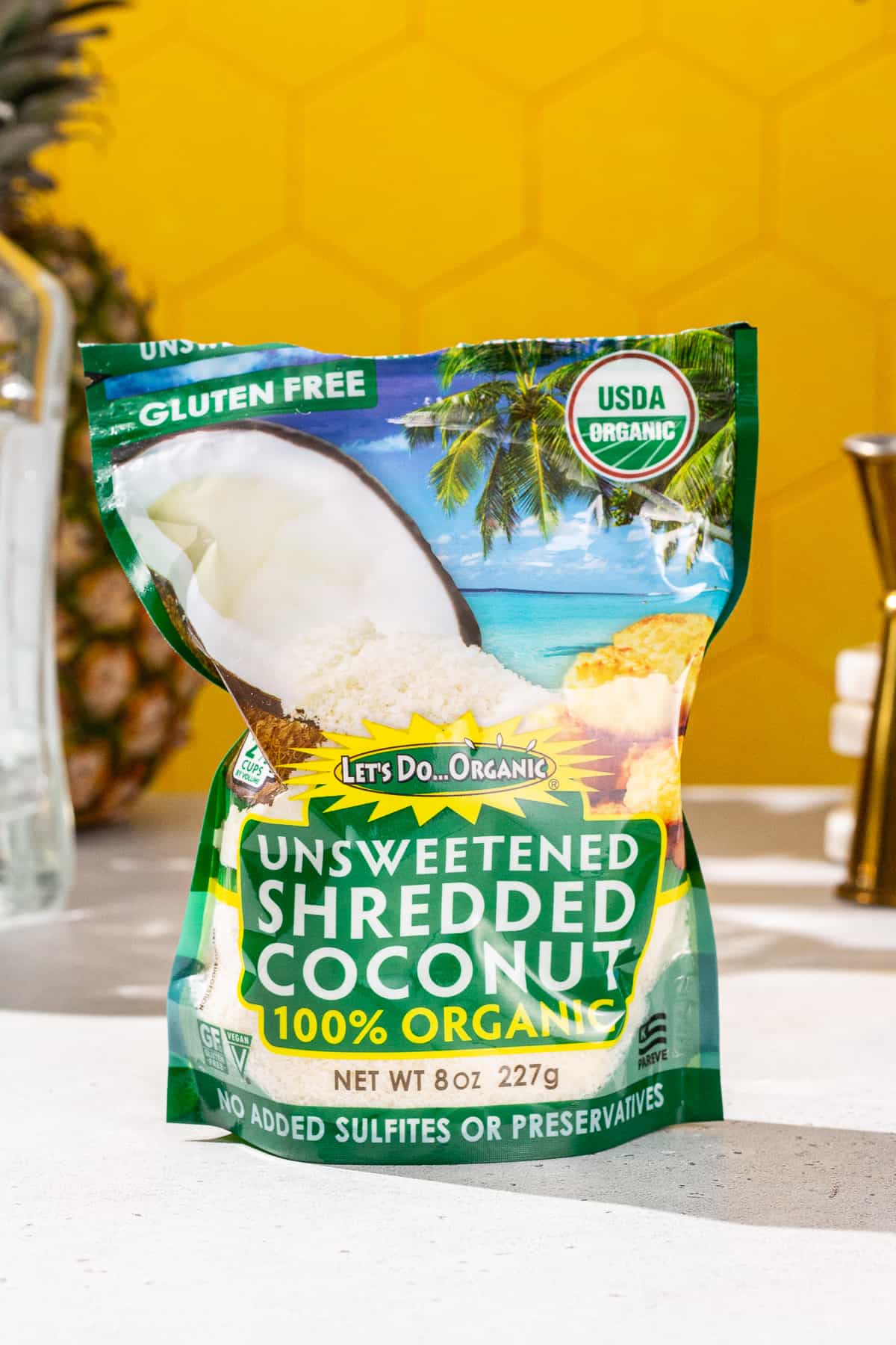 Bag of shredded coconut on a countertop, with a pineapple and tequila in the background.