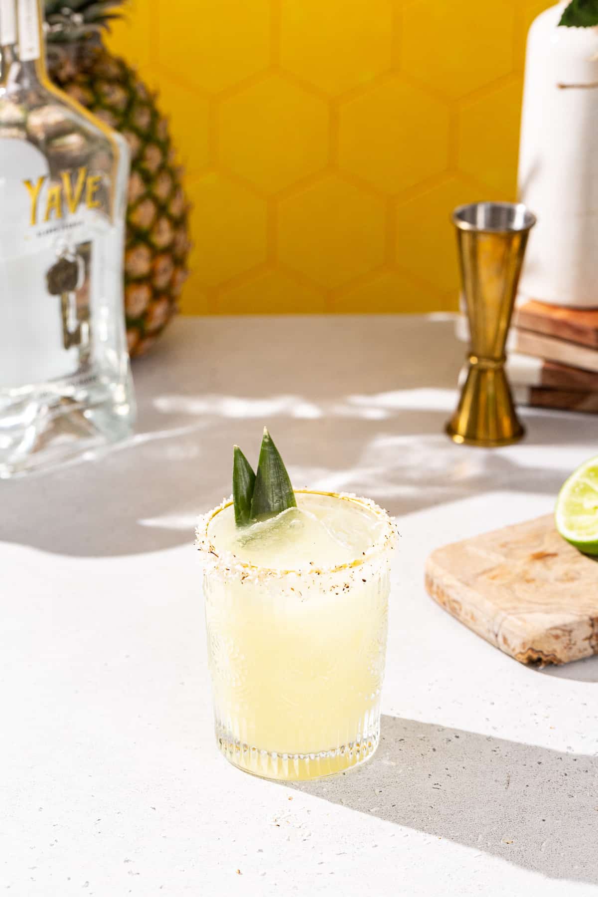 Slightly overhead view of a pineapple coconut margarita cocktail on a countertop. The glass is garnished with a toasted coconut rim and two pineapple fronds. Bar tools and ingredients are in the background.