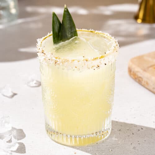 Side view of a pineapple coconut margarita cocktail on a countertop. The glass is garnished with a toasted coconut rim and two pineapple fronds. Some ice is on the counter next to the drink.