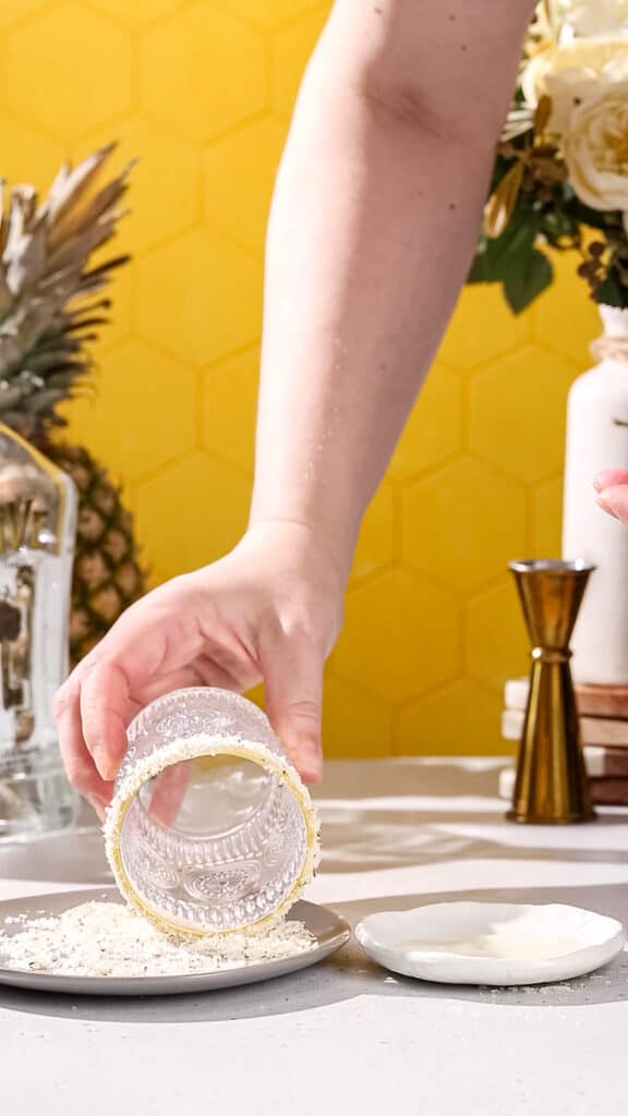 Hand dipping the rim of a cocktail glass into a dish of toasted dried shredded coconut.