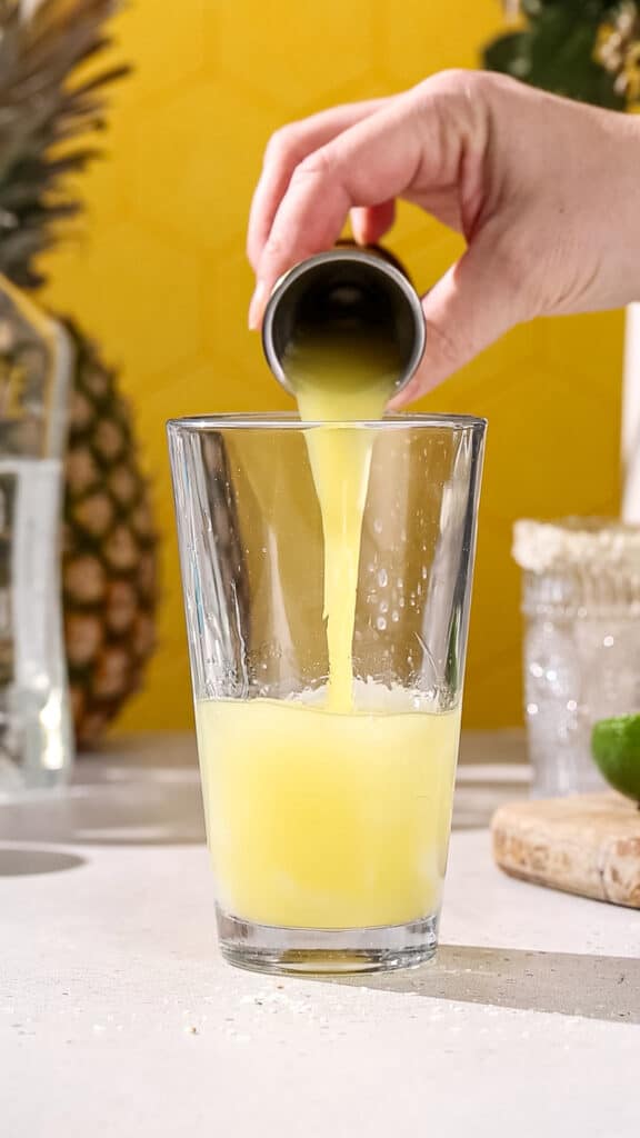 Hand pouring pineapple juice into a cocktail shaker.