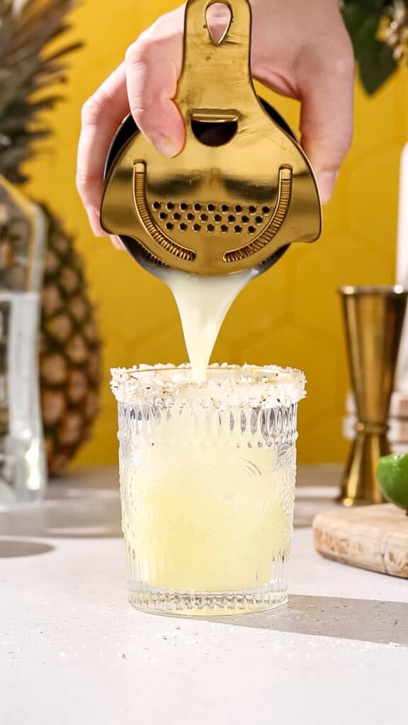 Hand straining a cocktail into a coconut-rimmed cocktail serving glass.