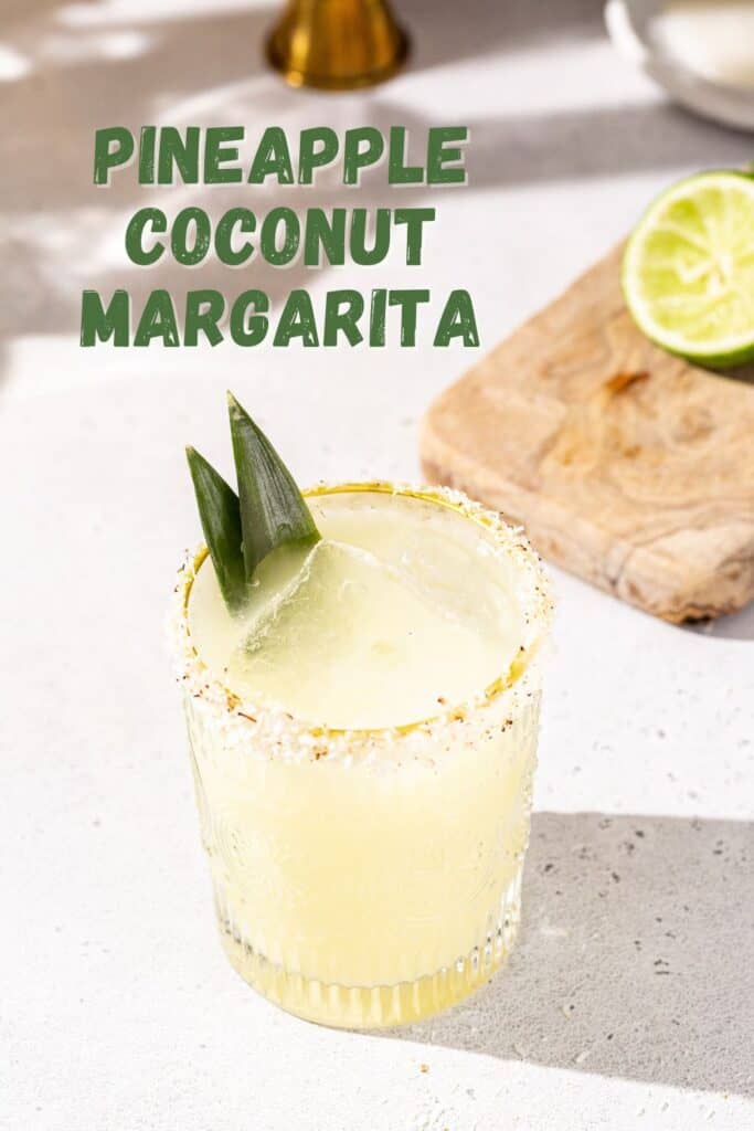 Overhead view of a pineapple coconut margarita cocktail on a countertop. The glass is garnished with a toasted coconut rim and two pineapple fronds. A cut lime is on a cutting board next to the drink. Text overlay says “Pineapple Coconut Margarita” in bold green letters.