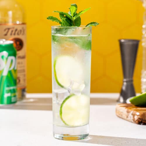Side view of a Vodka Press cocktail with fresh mint and lime garnish. In the background are bar tools and ingredients used to make the drink, including vodka and Sprite soda.
