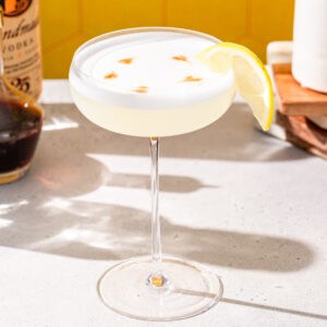 Vodka Sour cocktail on a countertop. The drink is in a tall stemmed cocktail glass and it is garnished with a half lemon slice as well as drops of bitters on top of the cocktail foam. Ingredients to make the drink are visible in the background.