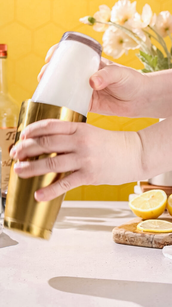 Hands shaking a cocktail in a gold and glass cocktail shaker filled with liquid and ice.