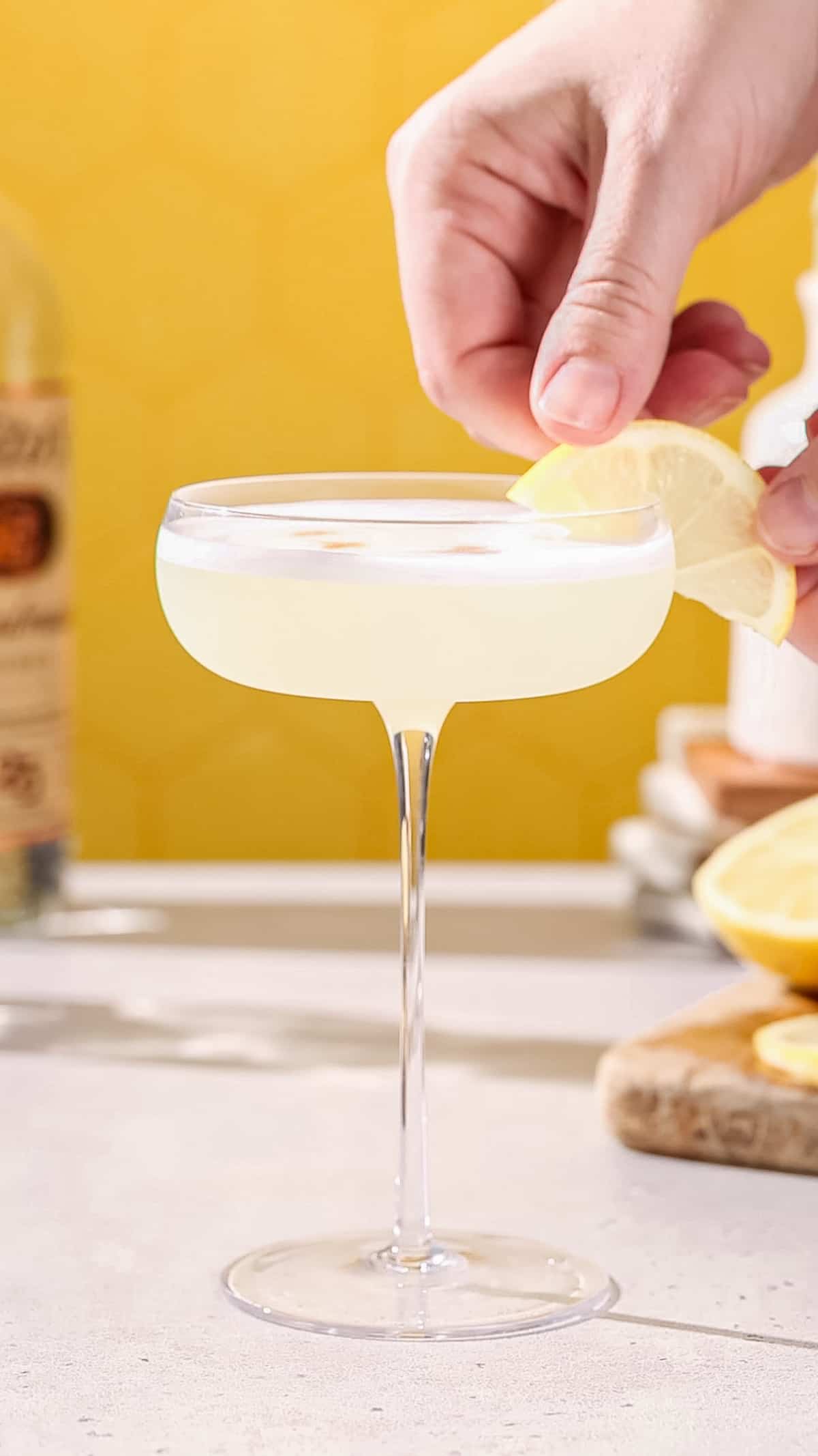 Hand adding a lemon slice to garnish a cocktail in a coupe glass.