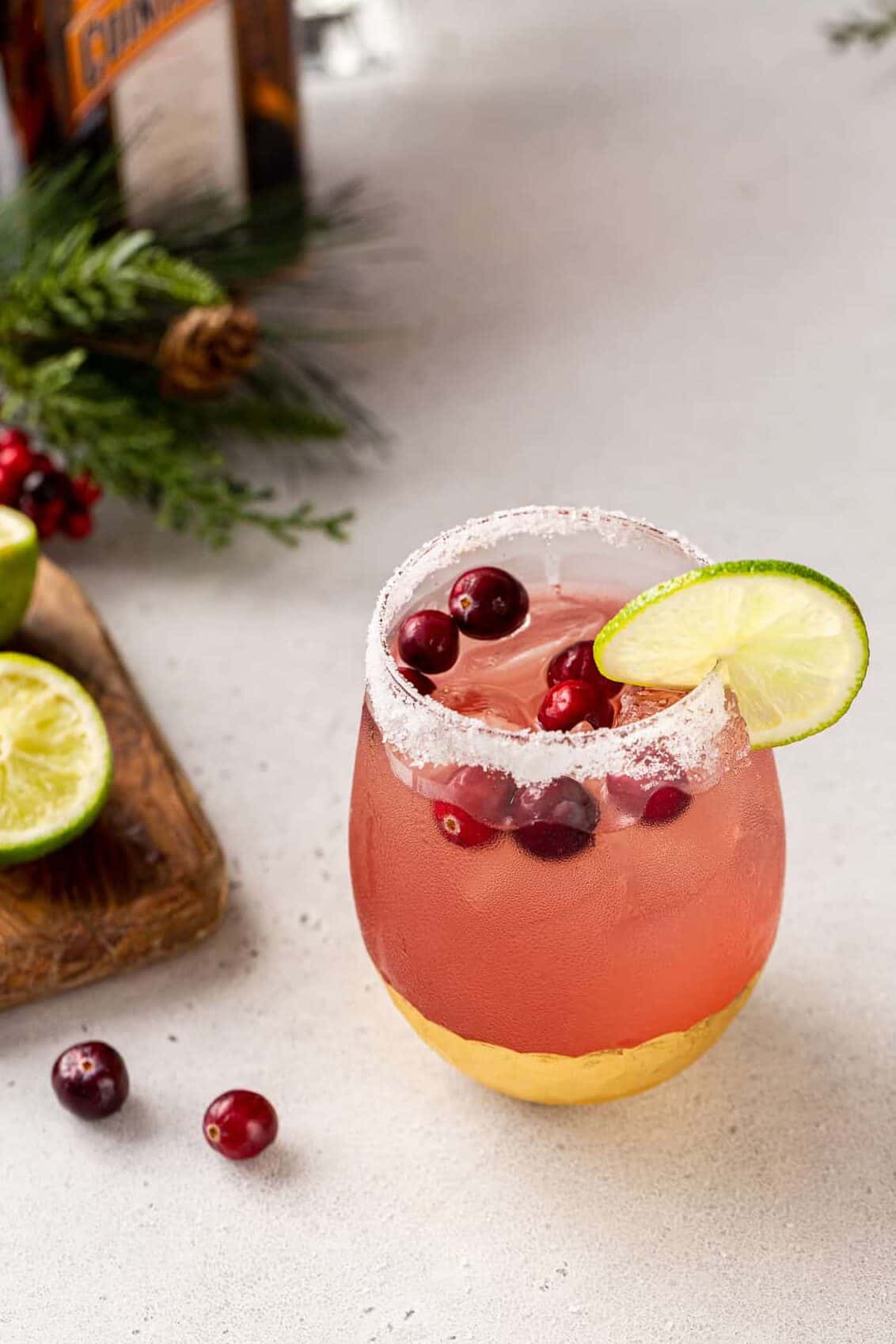A Cranberry Margarita on a gray countertop in a gold-bottomed glass. The margarita has a cut lime garnish and cranberries floating on top. To the left of the glass are cranberries, cut limes on a cutting board, pine bough decorations, and a bottle of Cointreau.