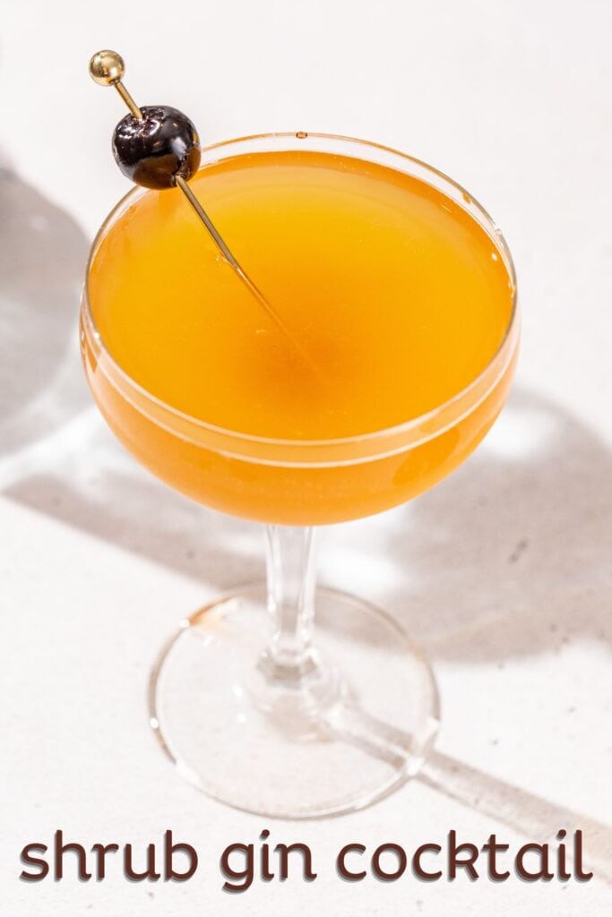 Overhead view of an orange colored cocktail in a coupe glass with a maraschino cherry garnish. Text overlay at the bottom says "shrub gin cocktail".