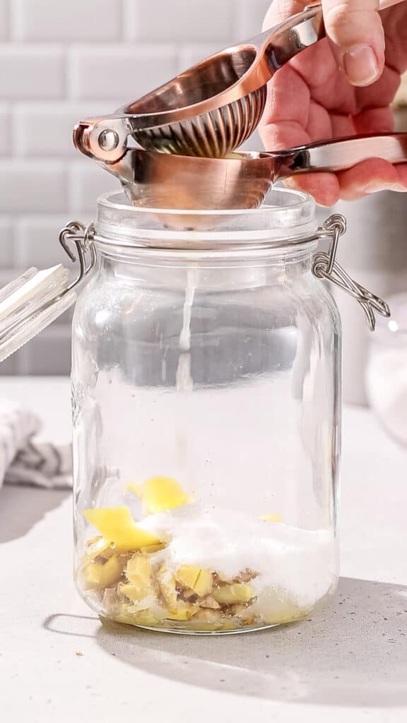 Hands using a citrus juicer to juice a lemon into a glass jar filled with ginger, lemon peels and sugar.