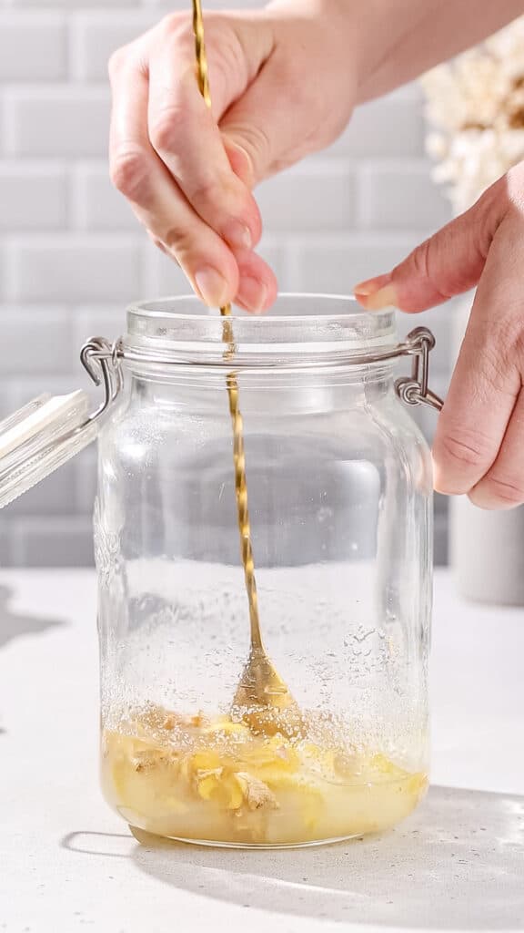 Hand using a spoon to stir a mixture of ginger, lemon and sugar in a glass jar.