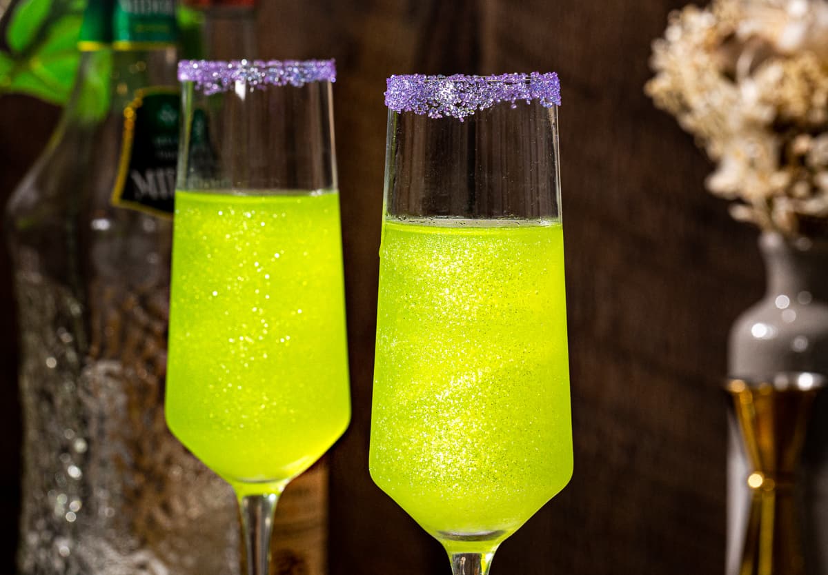 Side view of two Champagne flutes filled with bright green glittery cocktails. The rims are covered in purple sugar.