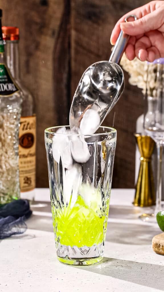 Hand using an ice scoop to add ice to a green cocktail in a cocktail shaker.
