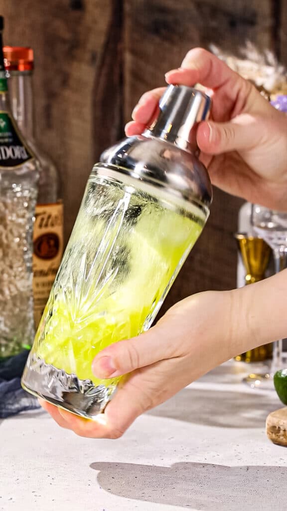 Hands shaking a green cocktail in a glass shaker.