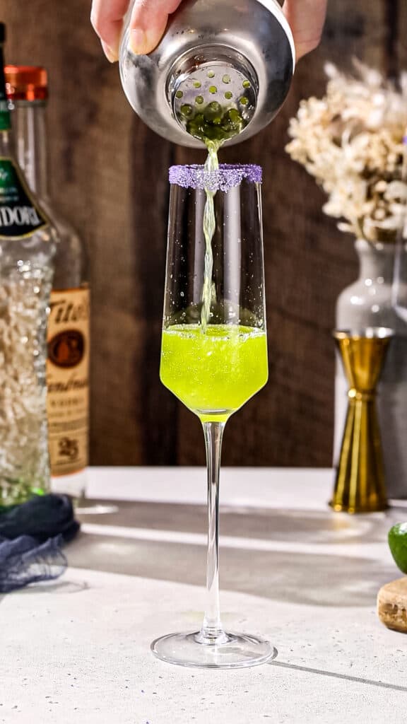 Hand using a cocktail shaker to strain a green drink into a Champagne flute.