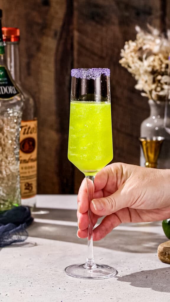 Hand about to pick up a Champagne flute filled with green sparkly liquid.