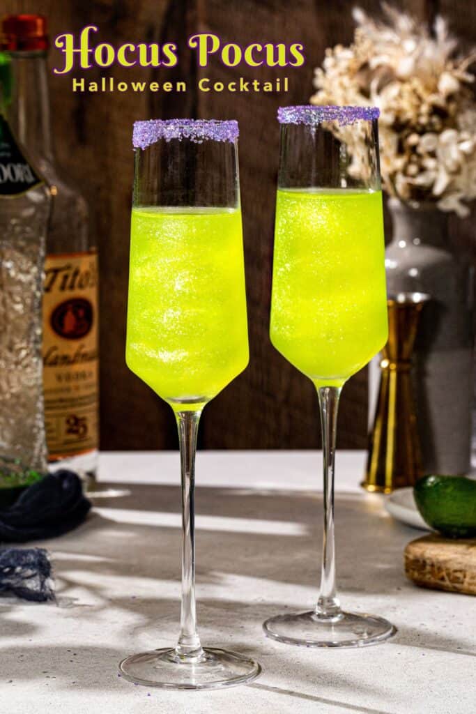 Side view of two green glittery Hocus Pocus cocktails with purple sugar rims on a countertop. Midori, vodka, a jigger and a lime are in the background. Text overlay at the top of the image says “Hocus Pocus halloween cocktail”.