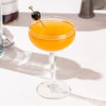 Slightly overhead view of a shrub cocktail in a stemmed glass. The cocktail is garnished with a maraschino cherry on a gold cocktail pick.